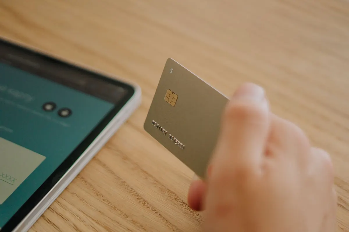 A hand holding a payment card beside a tablet showing a booking page
