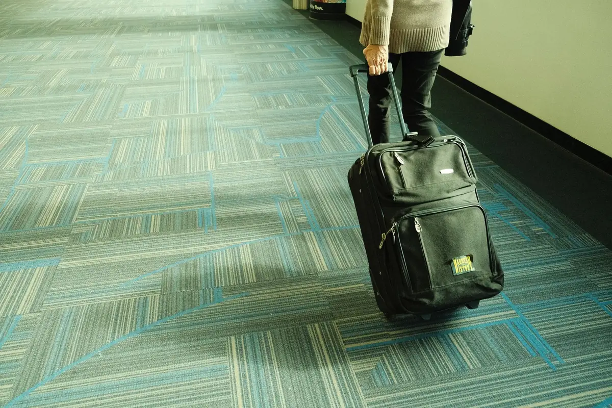 A traveler wheeling a suitcase down a long carpeted corridor, leaving for a trip