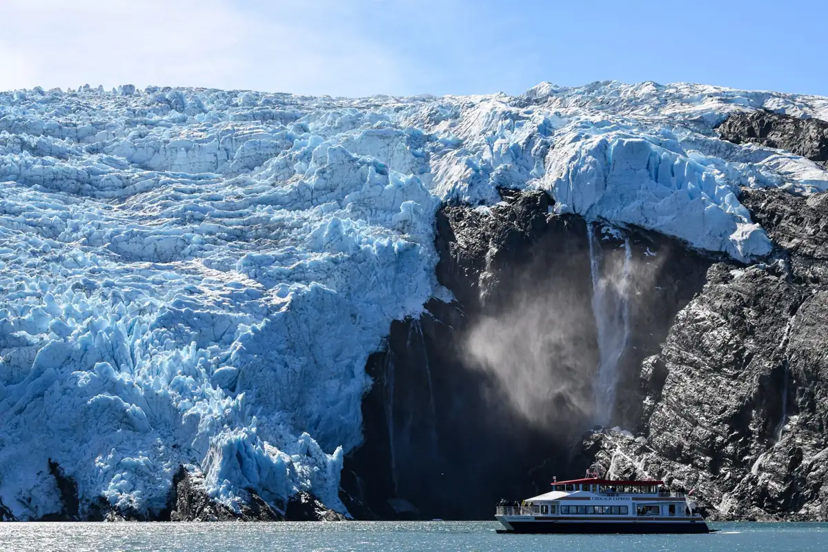 A blue glacier face with a waterfall beside it and a small tour boat below, in Alaska