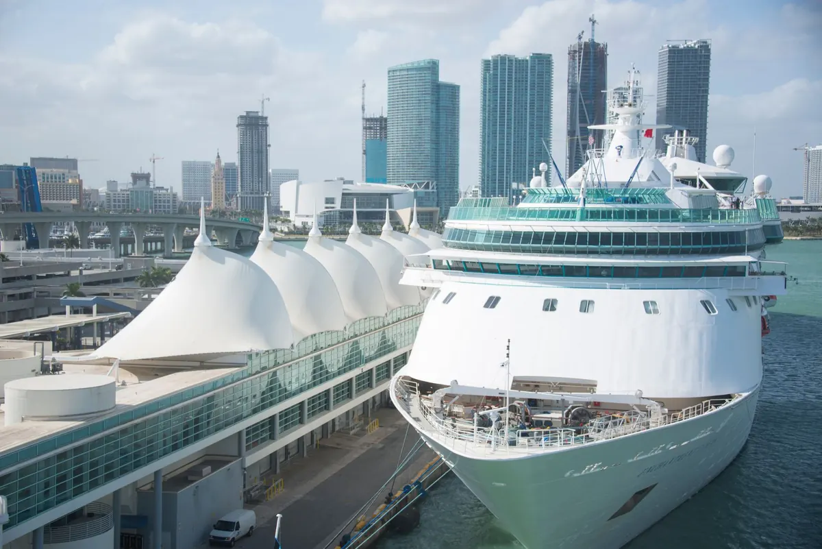 A white cruise ship at its berth in Miami, with the city skyline and the terminal roofs beside it