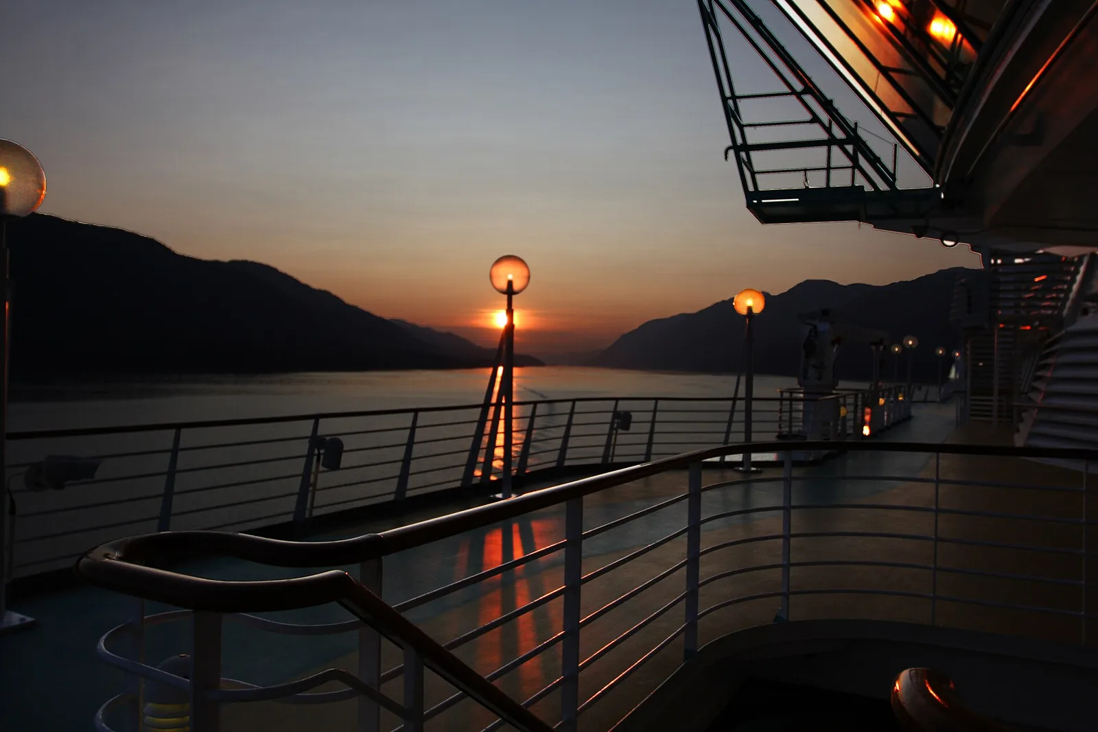 Sunset seen over the rail of a cruise ship deck, with mountains on both sides of the water