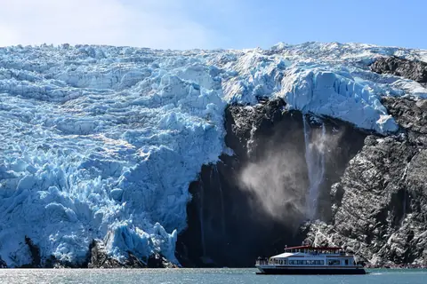 A blue glacier face with a waterfall beside it and a small tour boat below, in Alaska