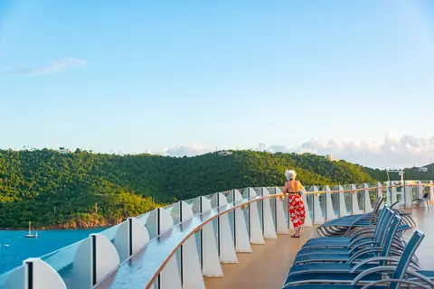 A traveler at the rail of a cruise ship deck looking out over green hills and blue water, the view a suite gives you