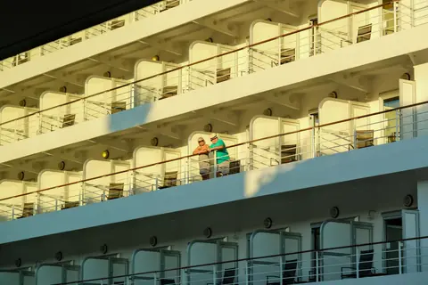 Two people standing on a cruise ship balcony in the sun, looking out
