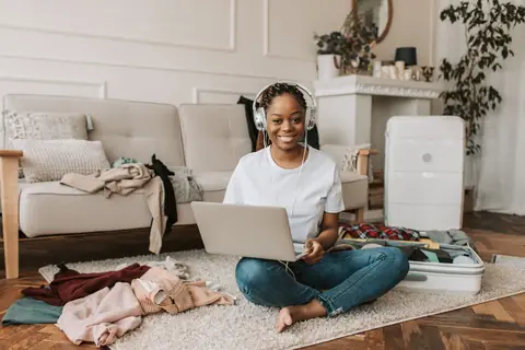 A smiling traveler on a living room floor with a laptop and a half-packed suitcase, checking fares before booking